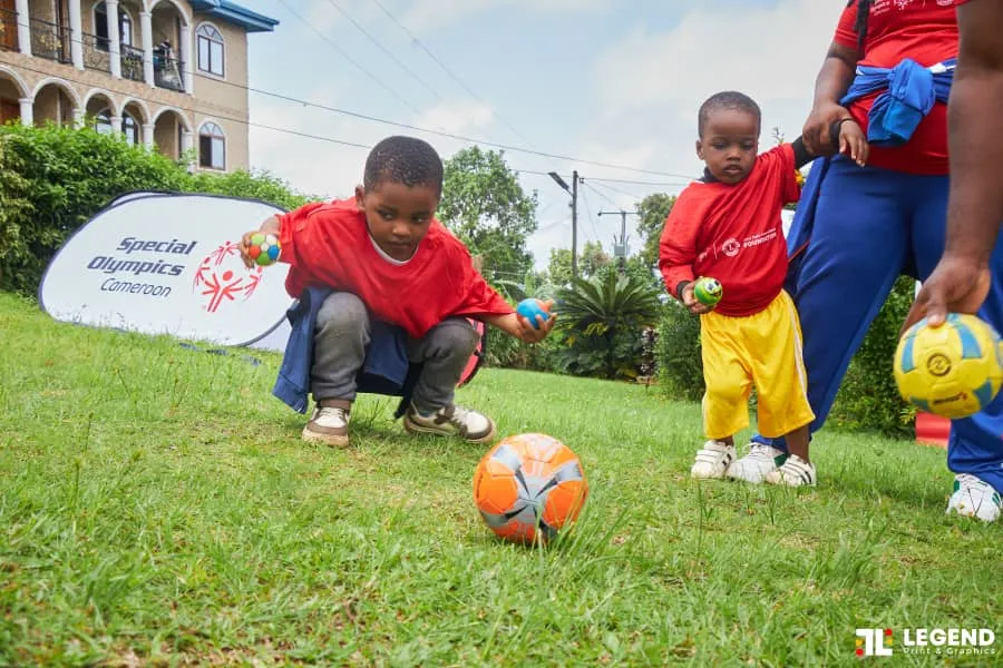 Deux jeunes enfants jouent avec des balles sur une pelouse devant une bannière de Special Olympics Cameroun