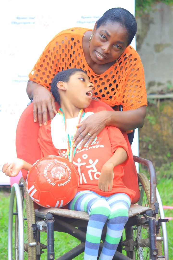 Une femme entoure de ses bras un enfant en fauteuil roulant, médaille au cou et ballon sur les genoux